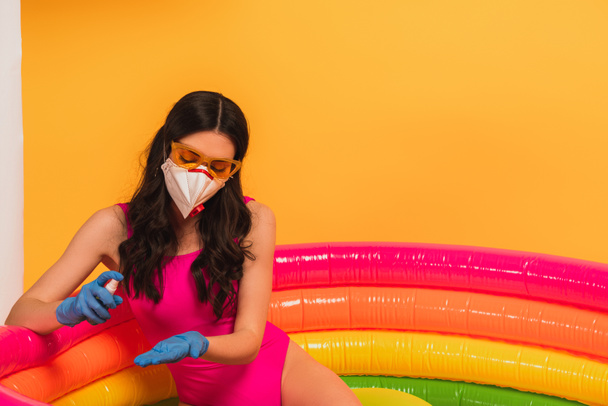 νεαρή γυναίκα με μαγιό, γυαλιά ηλίου, γάντια λάτεξ και ιατρική μάσκα κάθεται σε φουσκωτή πισίνα και εφαρμόζει απολυμαντικό στο κίτρινο - Φωτογραφία, εικόνα
