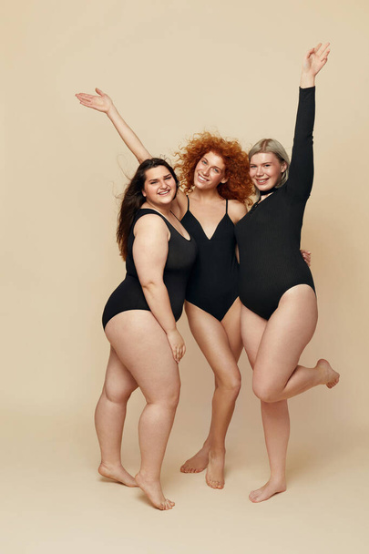 Different Body Types. Group Of Diversity Models Full-length Portrait. Blonde, Brunette And Redhead In Black Bodysuits Posing On Beige Background. Female Friendship For Happy Life.  - Fotó, kép