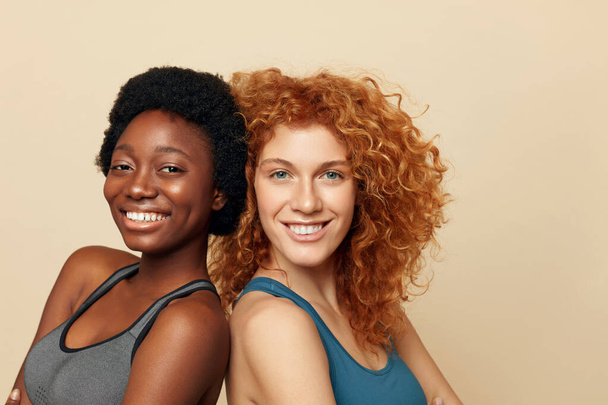 Fitness Women. Ethnic Friends Close Up Portrait. Smiling Brunette And Redhead Posing On Beige Background. Sport As Lifestyle.  - 写真・画像