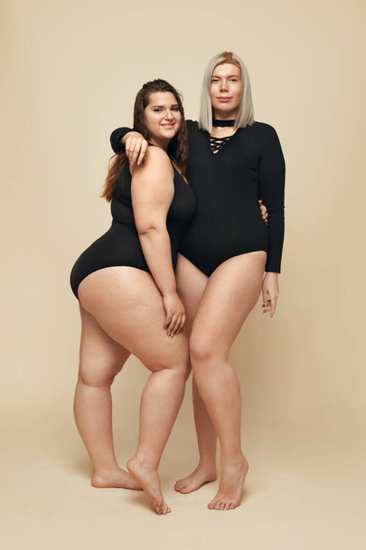 Plus Size Models. Full-figured Women Full-length Portrait. Brunette And Blonde In Black Bodysuits Posing On Beige Background. Body Positive Concept.  - 写真・画像