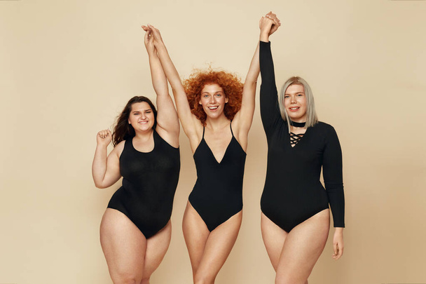 Different Body Types. Group Of Diversity Models Portrait. Smiling Blonde, Brunette And Redhead In Black Bodysuits Holding Hands Up. Female Friendship For Happy Life.  - Fotó, kép