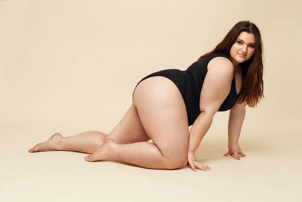Plus Size Model. Fat Woman In Black Bodysuit Portrait. Crawling Brunette Posing On Beige Background And Looking At Camera. Body Positive Concept.  - Foto, afbeelding