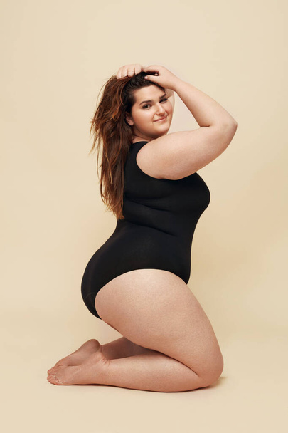 Plus Size Model. Fat Woman In Black Bodysuit Portrait. Brunette Touching Hair And Posing On Beige Background. Body Positive Concept.  - Zdjęcie, obraz
