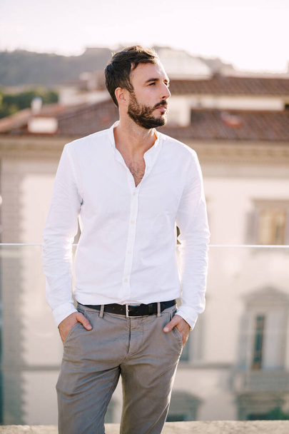 Stílusos szakállas fickó fehér ingben és könnyű nadrágban egy tetőteraszon Firenzében, Olaszországban - Fotó, kép