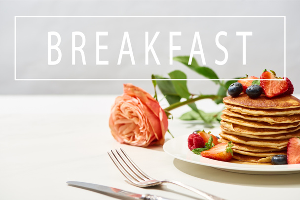 selectieve focus van heerlijke pannenkoeken met bosbessen en aardbeien op bord in de buurt van rozenbloem en bestek op wit oppervlak geïsoleerd op grijs, ontbijt illustratie - Foto, afbeelding