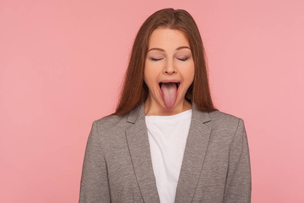Portrait of carefree young woman in business suit sticking out tongue and keeping eyes closed, teasing with naughty expression, disobedient behavior. indoor studio shot isolated on pink background - 写真・画像