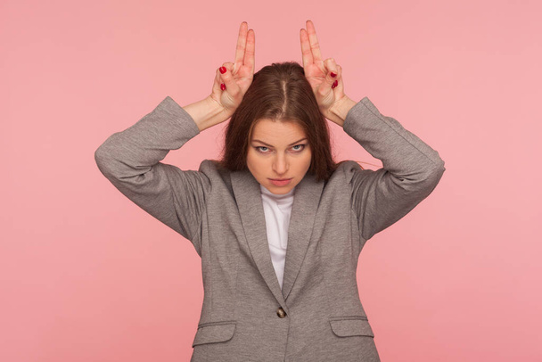 I am dangerous! Portrait of aggressive young woman in business suit standing with bull horn hand sign, antler gesture on head, threatening to attack enemy. studio shot isolated on pink background - Foto, afbeelding