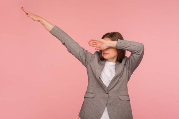 Portrait of successful young woman in business suit showing dab dance pose, famous internet meme of triumph, performing dabbing trends with hand gesture. studio shot isolated on pink background - Photo, Image