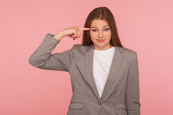 This is insane idea! Portrait of woman in business suit showing stupid gesture with finger at her temple, displeased with reckless silly joke, out of mind. studio shot isolated on pink background - Photo, image