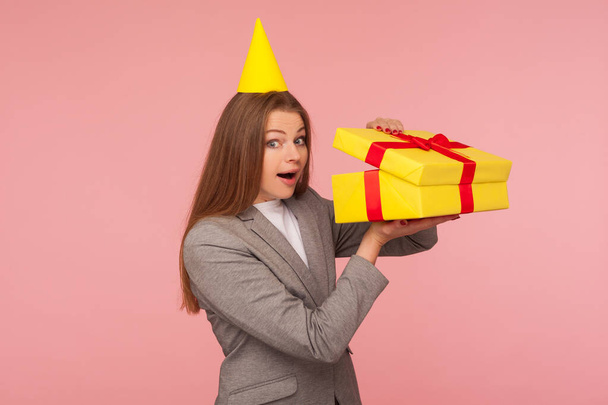 Portrait of funny nosy woman in business suit and with party cone on head unboxing present and looking with amazement, astonished by gift, bonus, unwrapping birthday surprise. studio shot isolated - 写真・画像