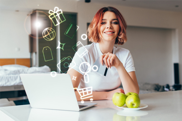 smiling girl shopping online with laptop and credit cards and shopping signs on kitchen with apples during self isolation  - Фото, изображение