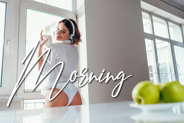 happy girl listening music in headphones and holding coffee cup on kitchen with apples during self isolation with morning lettering - 写真・画像