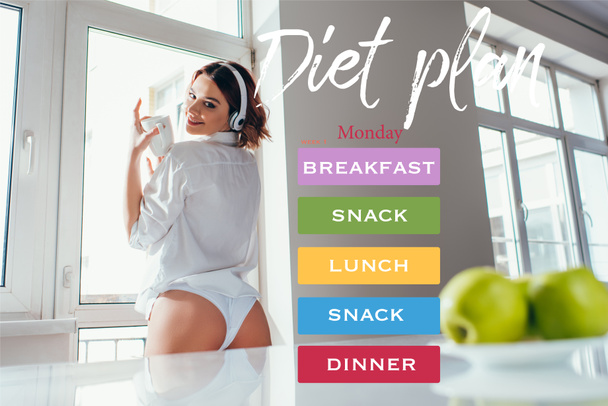 happy girl listening music in headphones and holding coffee cup on kitchen with apples during self isolation with diet plan lettering - Photo, Image