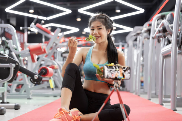 Spor salonunda sebze yiyen sağlıklı Asyalı genç bir kadın ve blog yazarı vlog için video kaydediyor. Fitness ve sağlıklı yaşam tarzı konsepti - Fotoğraf, Görsel