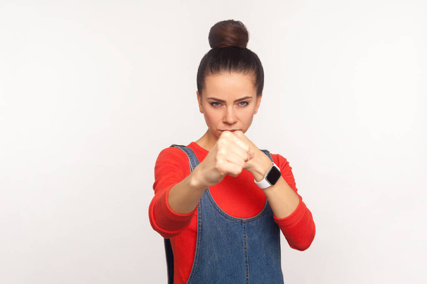 Let's fight! Portrait of determined aggressive confident girl with hair bun in denim overalls boxing with clenched fist, punching in front of camera, struggle and self-defence. studio shot, isolate - Фото, зображення