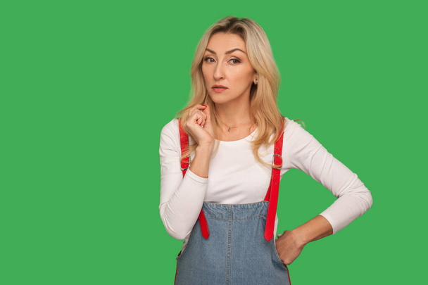 Portrait of pensive concerned adult blond woman in stylish denim overalls looking thoughtful and confused by dilemma, pondering idea with doubtful suspicious expression. indoor studio shoot isolated - Zdjęcie, obraz