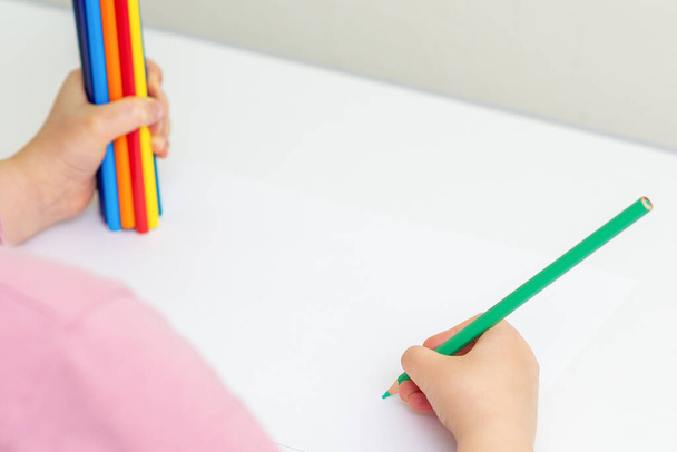 Ребёнок рисует зелёным карандашом на белой бумаге, держа в руке цветные карандаши. Селективный фокус
. - Фото, изображение