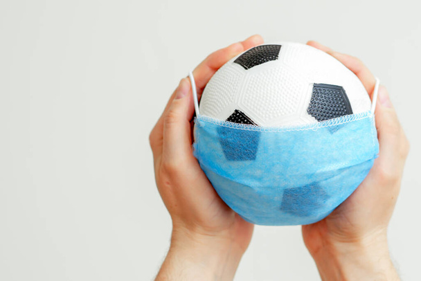 Closeup of hands of man holding soccer ball in medical mask on a light background with copy space. Cancellation of sporting events. - Foto, Imagen