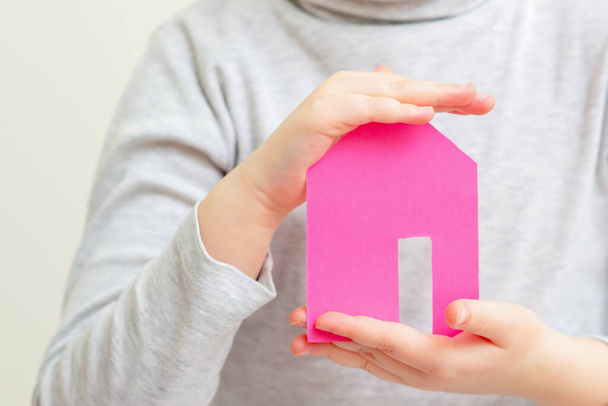 Gros plan des mains de la petite fille tenant la maison en papier rose souriant et rêvant. Concept de famille et de maison
. - Photo, image