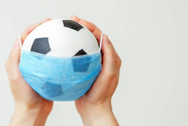 Closeup of man hands holding soccer ball in medical mask on light background during coronavirus. Football paused because coronavirus. - Fotografie, Obrázek