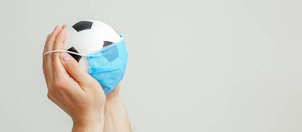 Hands of man holding soccer ball with a medical mask on a gray background with copy space. Quarantined football. - Foto, afbeelding