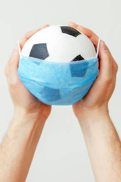 Closeup vertical image of soccer ball with medical protective mask in hands of man on light background during coronavirus. Cancellation of football competitions. - Zdjęcie, obraz