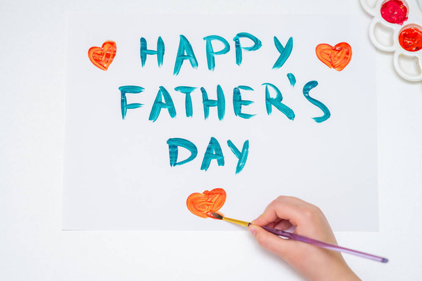Зверху погляд на дитячу руку, що малює червоне серце з текстом "Щасливий день батька" на білому папері. З Днем батька.. - Фото, зображення