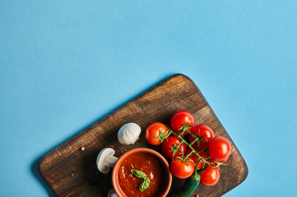 top view of delicious tomato sauce in bowl on wooden board with fresh ripe vegetables on blue background - Fotografie, Obrázek