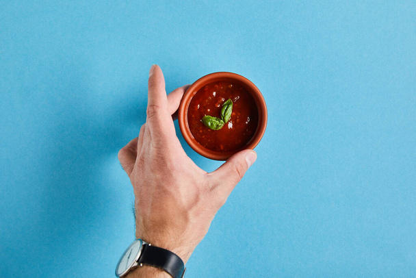 bovenaanzicht van mannelijke hand met heerlijke tomatensaus in schaal op blauwe achtergrond - Foto, afbeelding