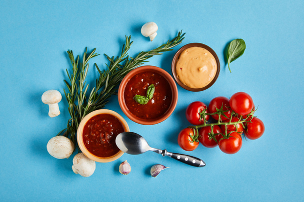 вид на вкусные соусы в мисках с ложкой рядом со свежими зрелыми овощами на синем фоне
 - Фото, изображение
