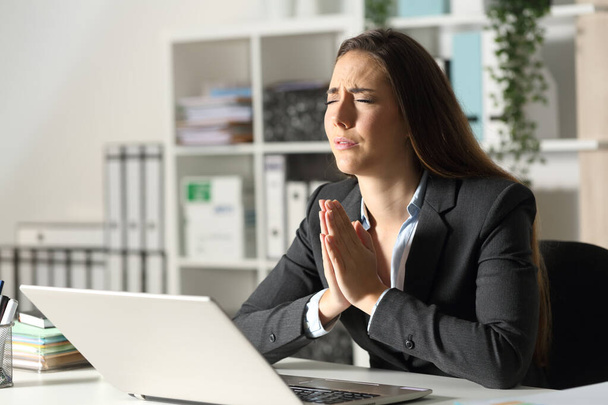 Worried executive woman with laptop praying sitting on her desk at night at office - Foto, Imagem