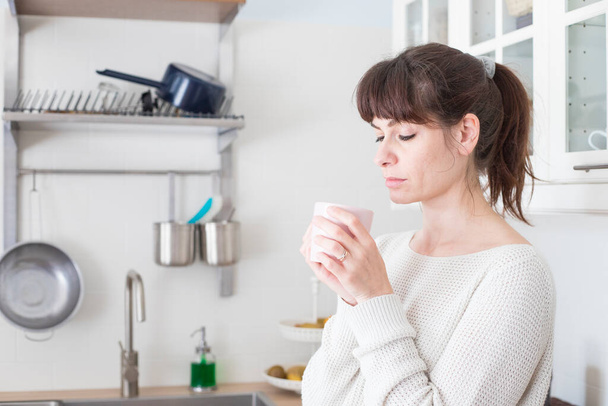Досить біла жінка, з чубчиком і хвостом, тримає чашку кави вранці в білому, яскравому освітленій кухні. Портрет профілю
. - Фото, зображення