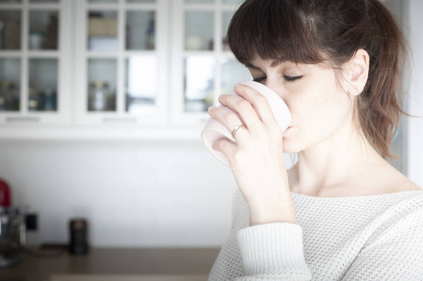 Pretty caucasian woman, with bangs and ponytail, drinking a cup of coffee in the morning in a white, bright lit, kitchen. Copy space. - Photo, image