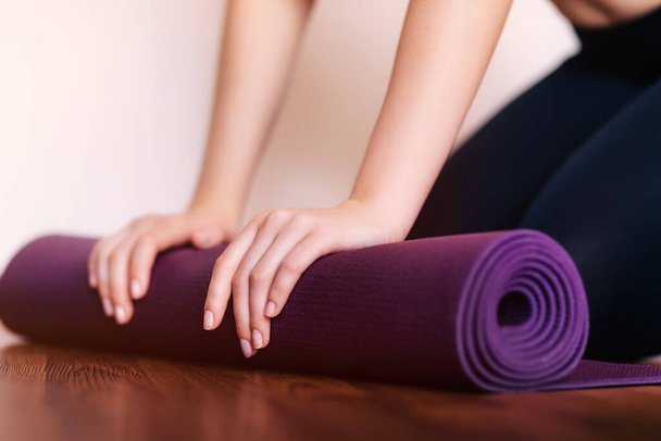 Gros plan de la femme pliant yoga violet ou tapis de fitness après l'entraînement à la maison dans le salon. Mode de vie sain, garder la forme concept. - Photo, image