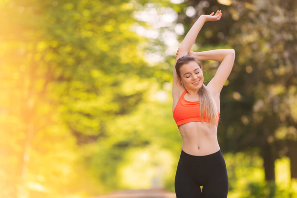 Young slim woman with a sporty body, long hair, dressed in a sports pink top and leggings, prepares for a morning jog in the park. Copy space - Zdjęcie, obraz