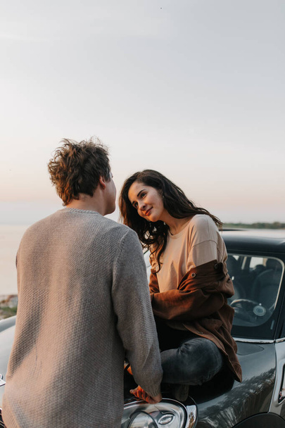 Beautiful young couple in love kisses and hugs sitting on a car - Photo, image
