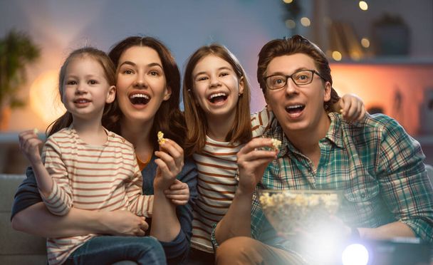 Happy family watching projector, TV, movies with popcorn in the evening at home. Mother, father and daughters spending time together. - Foto, afbeelding