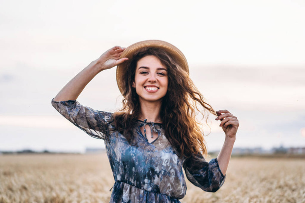 Closeup portrait of a beautiful young woman with curly hair. Woman in dress and hat standing in wheat field. - Foto, Bild