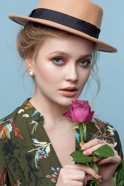 Portrait of attractive smiling woman blond isolated on blue studio background with pink rose near lips. Close up portrait, fashion look in a hat. - Foto, immagini