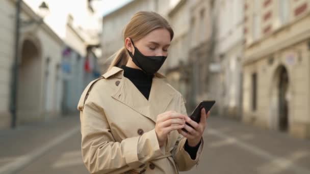 Egy fiatal szőke nő orvosi maszkban sétál az utcán. telefonos üzeneteket használ tekercsek szörfözik az interneten. keresési hírek covid19 coronavirus virus protection pandemic city. - Felvétel, videó