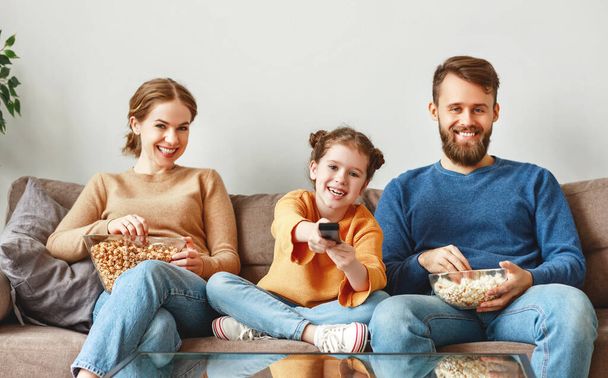 Optymistyczna rodzina w luźnym ubraniu, siedząca na kanapie w świetlicy i śmiejąca się podczas oglądania telewizji i jedzenia smacznego popcornu w słoneczny dzień w Hom - Zdjęcie, obraz