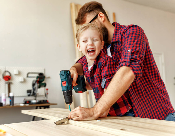 Muchacho emocionado y padre joven en camisas similares que sostienen taladro juntos mientras se trabaja con madera en los estudios de artesanía moderna
 - Foto, imagen
