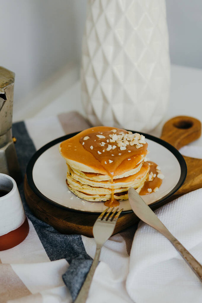 Pfannkuchen mit gesalzenem Karamell und Erdnüssen neben Kaffeemaschine auf weißem Tisch in der Küche. Morgens Frühstück zu Hause in Quarantäne. Rezept von hausgemachten leckeren Pfannkuchen. - Foto, Bild