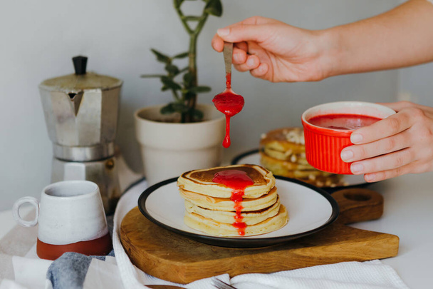 Girl pouring raspberry sauce on pancakes near coffee maker on white table in kitchen. Morning breakfast at home in quarantine. Recipe of homemade delicious pancakes. - Valokuva, kuva