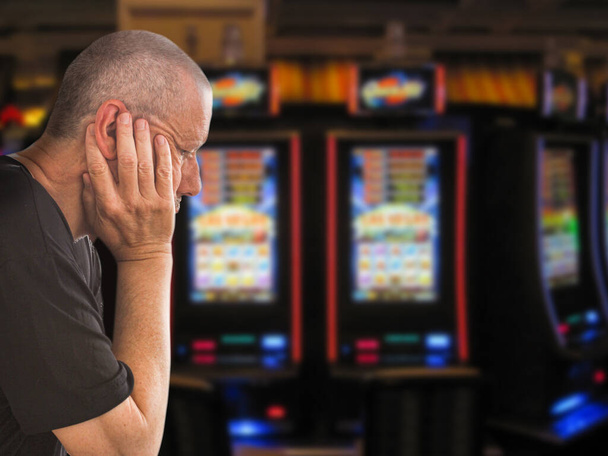 Sad and depressed caucasian man sitting with his hands on his head in front of rows of casino slot machines. Gambling addiction theme image.  Close up portrait. - Foto, afbeelding