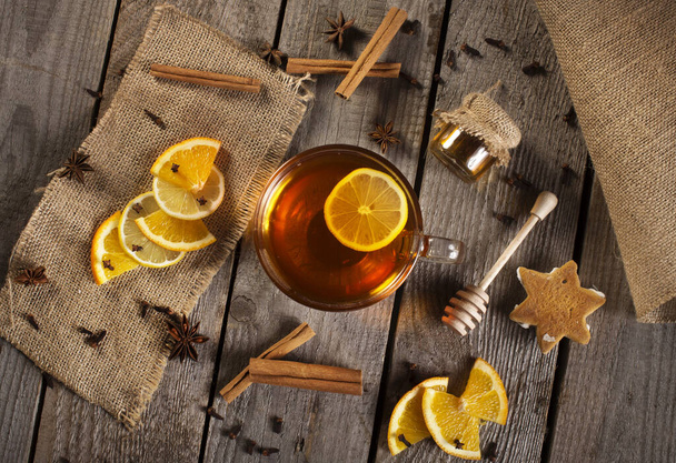 Cup of hot tea on rustic wooden plank with cinnamon stick, orange, lemon slices, honey, anise, cloves and other decoration. Vintage flat lay. - Photo, image