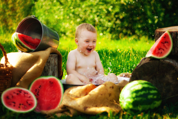 Retrato de un niño sin camiseta en el pueblo, al aire libre. Dos años de edad, niño comiendo pedazo de sandía en la hierba en el jardín. Cara sucia, niño feliz y emocional, sandía jugosa
. - Foto, imagen
