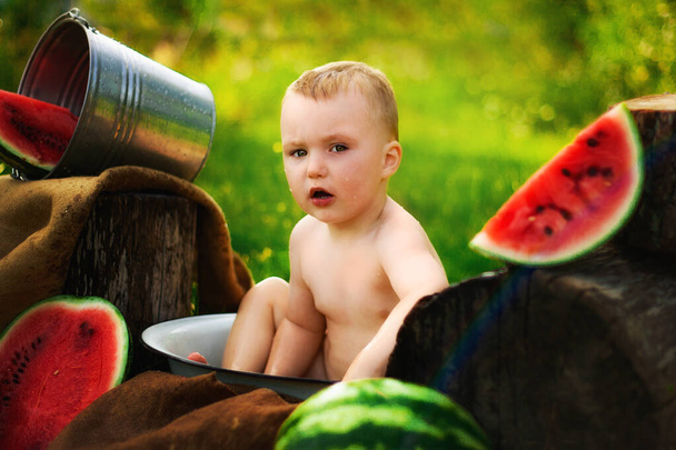 Πορτρέτο του μικρού παιδιού χωρίς t-shirt στο χωριό, στον καθαρό αέρα. Δύο ετών παιδί τρώει κομμάτι καρπουζιού στο γρασίδι στον κήπο. Βρώμικο πρόσωπο, χαρούμενο και συναισθηματικό παιδί, ζουμερό καρπούζι. - Φωτογραφία, εικόνα
