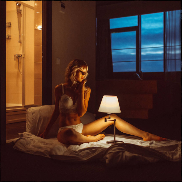 young beautiful blonde girl, evening in the hotel room alone in white lingerie and half-naked under the light from the lamp - Foto, Imagem