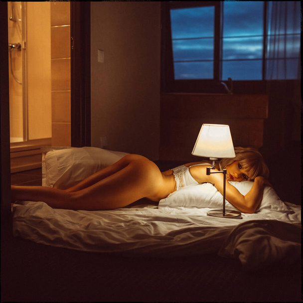 jovem linda menina loira, à noite no quarto de hotel sozinho em lingerie branca e semi-nua sob a luz da lâmpada
 - Foto, Imagem